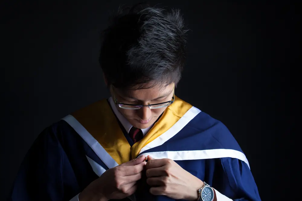 A graduate fastens his gown in low side light.