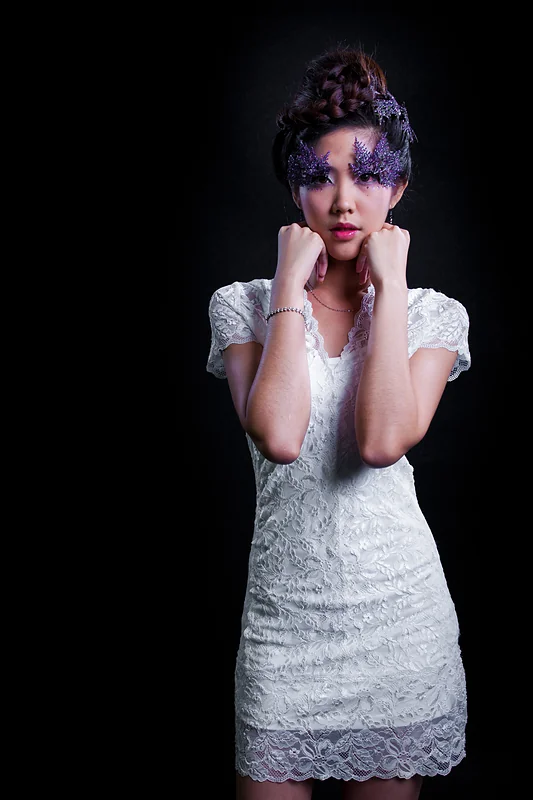 A woman in a white lace dress with purple eye make-up, lit against black.