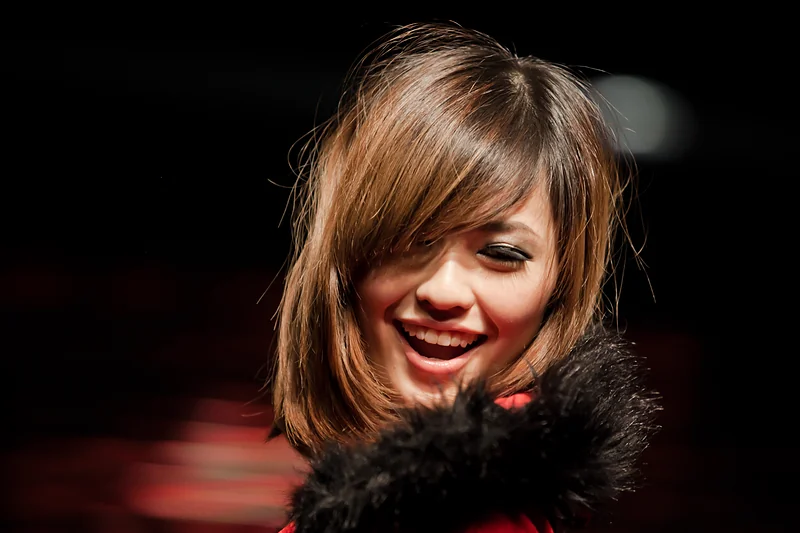 A woman laughs with her hair across her face, lit against a black background.