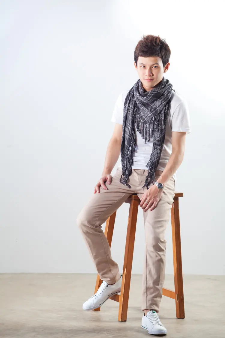 A young man in a checked scarf sits on a wooden stool in a white studio.
