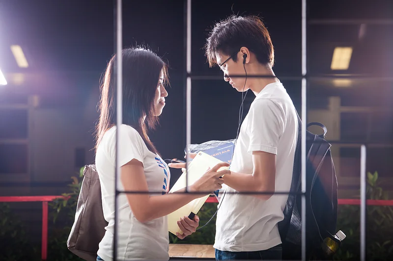 Two young people look at notes behind a window frame, in purple evening light.