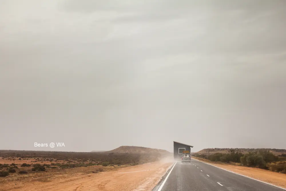 A truck comes out of a dust haze on a long, empty road.