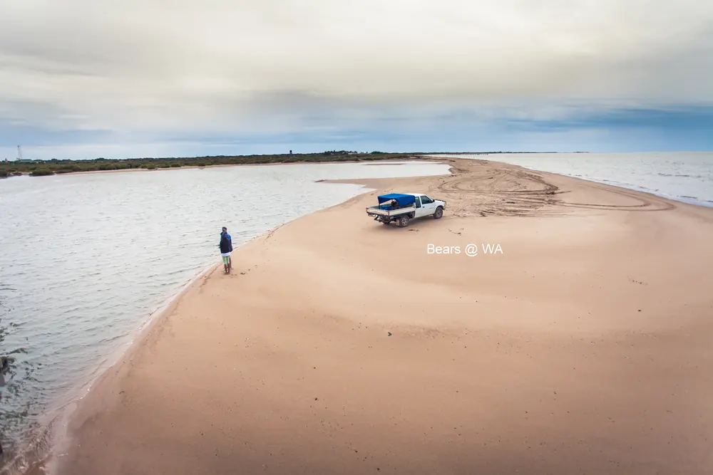 A person and a pickup truck on a long sand spit between two stretches of water.