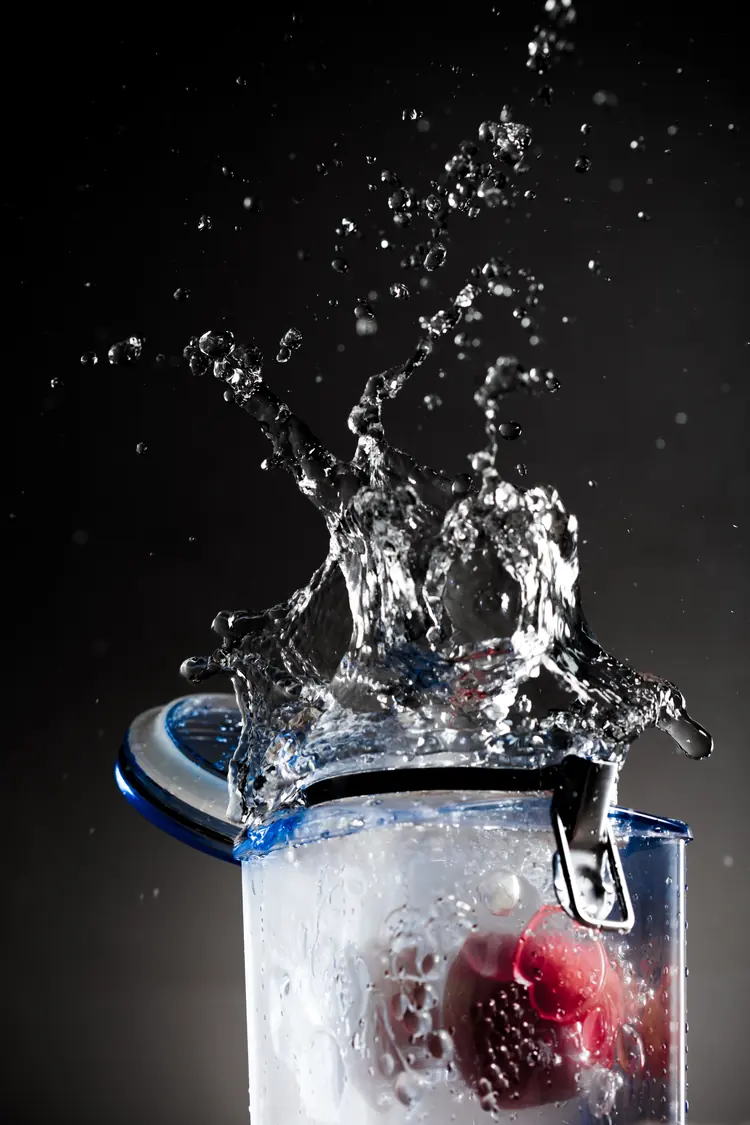 Water splashes out of a glass jar with a red fruit inside, against black.